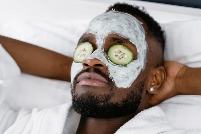 A face shot of a relaxing man in a robe with hands behind his head while lying down with a all-natural non-toxic face moisturizing mask on and cucumbers over his eyes.