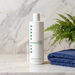 Natural Shampoo for Dry scalps bottle by Happy Lad on a marble surface with a blue towel and plant.