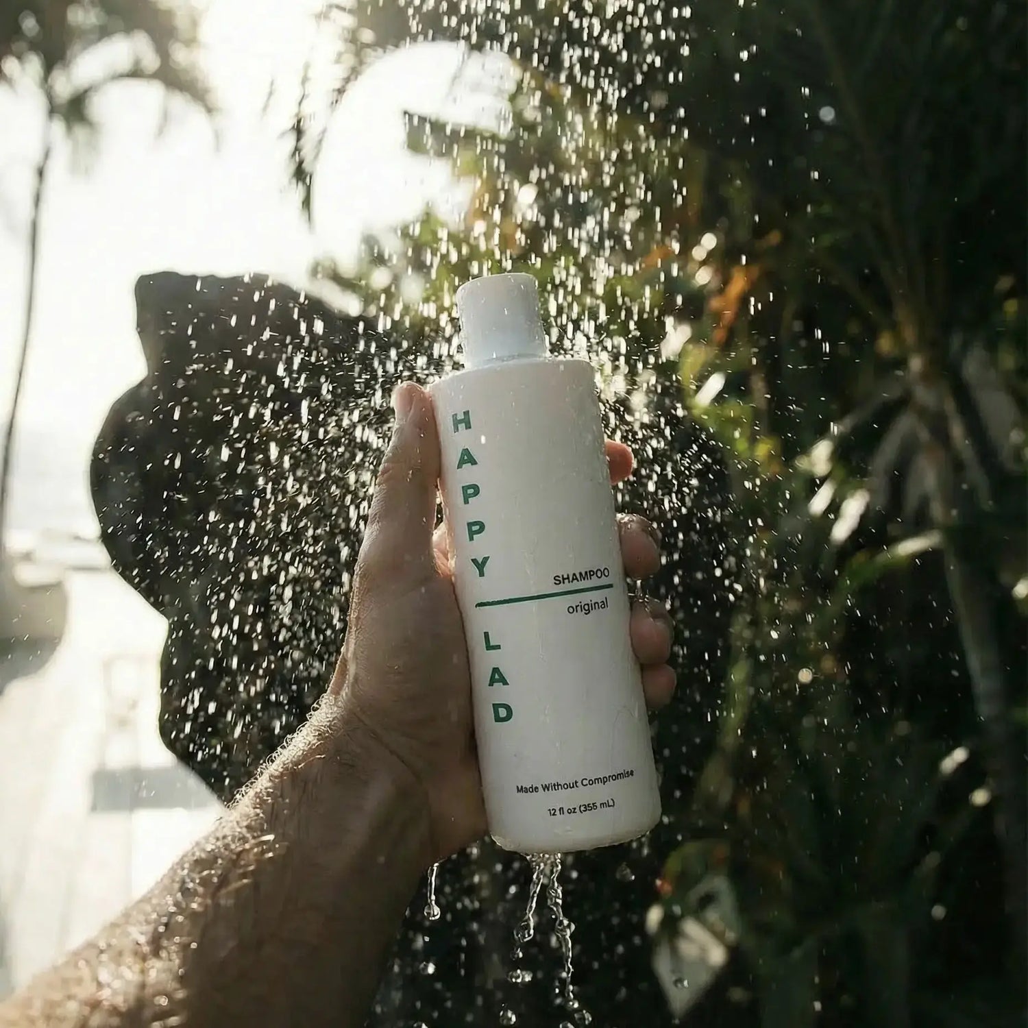 Hand holding a bottle of 'Happy Lad' anti itch natural shampoo under outdoor shower as water splashes with plants in the background