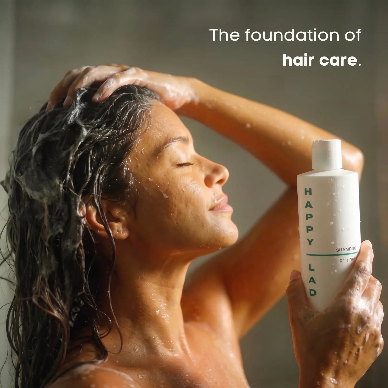 A woman with wet, soapy hair holds a natural shampoo by Happy lad. Text on the image reads 'The foundation of hair care.'